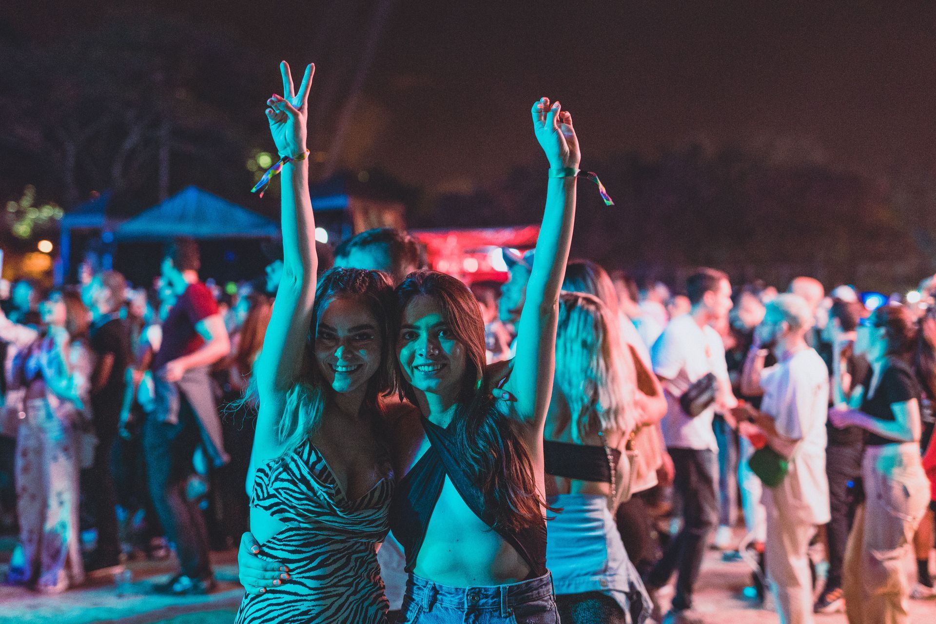 Two women dancing at a colorful nighttime outdoor party, with a crowd in the background