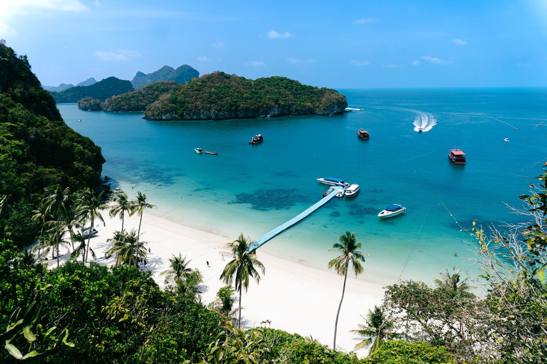 Tropical bay with turquoise water, white beach, boats, and a long pier under a clear blue sky