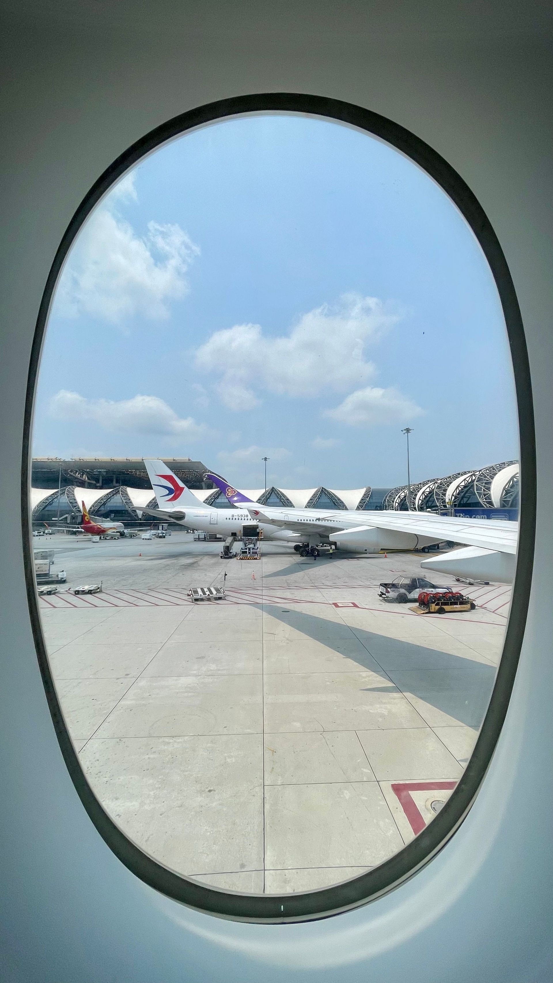 Airport runway seen through oval airplane window, with parked jets and a blue sky.