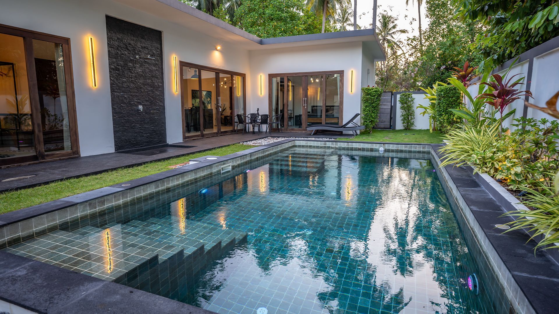 Modern home patio with a rectangular swimming pool, glass doors, and lush tropical plants at dusk