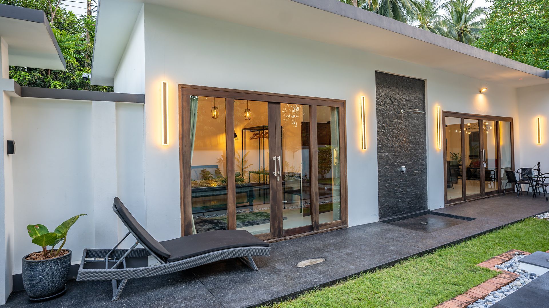 Modern white house patio with glass doors, lounge chairs, warm wall lights, and a small lawn