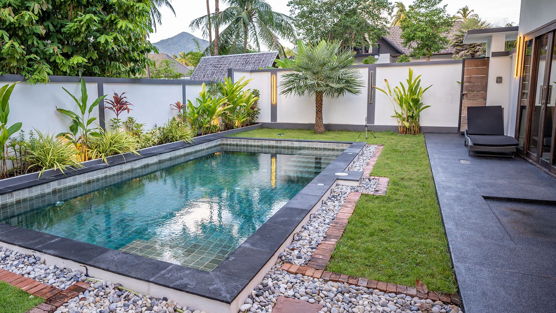 Backyard pool with stone edging, green lawn, tropical plants, and a lounge chair beside a white wall