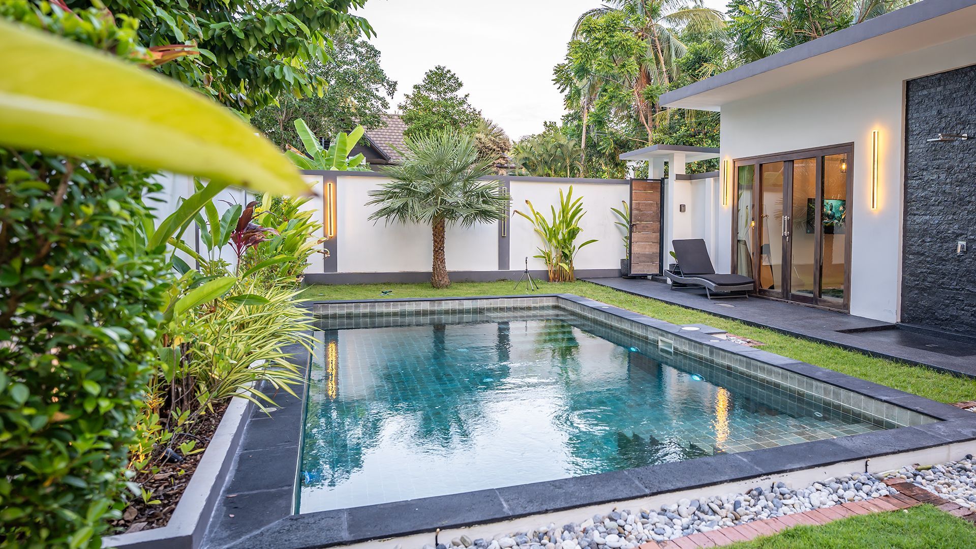 Modern backyard pool beside a house, with tropical plants and lounge chairs in a landscaped patio area