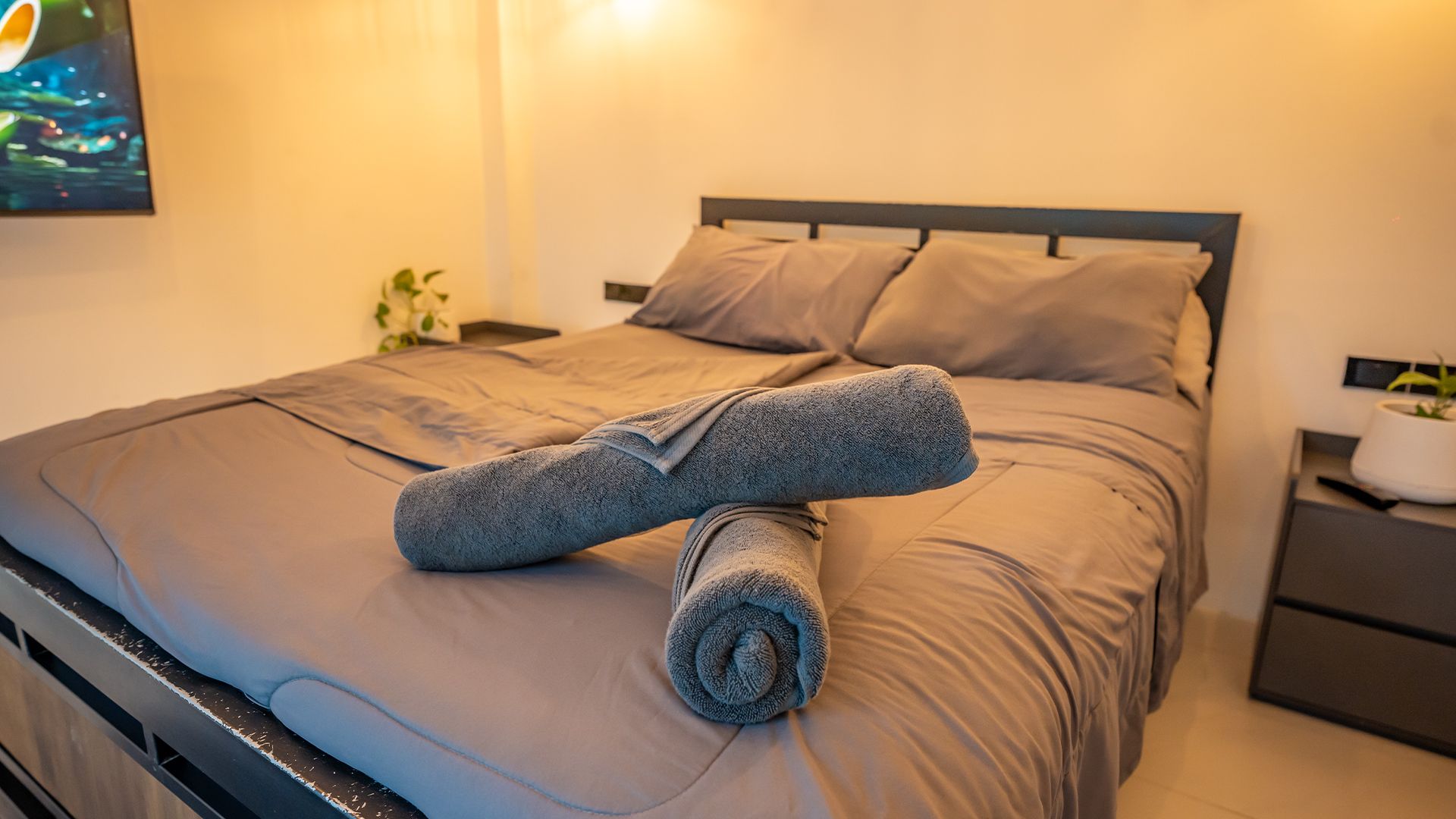Neatly made bed with gray bedding and rolled towels in a warm, softly lit bedroom.