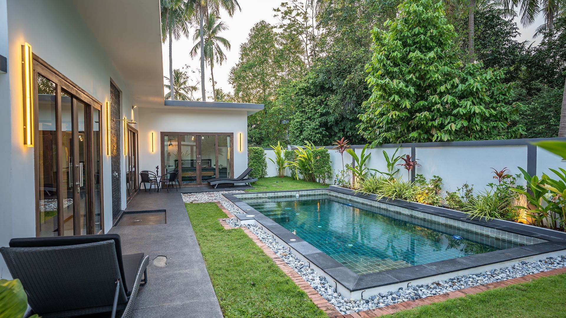 Modern backyard with a rectangular pool, lounge chairs, and a covered patio beside a white wall and greenery