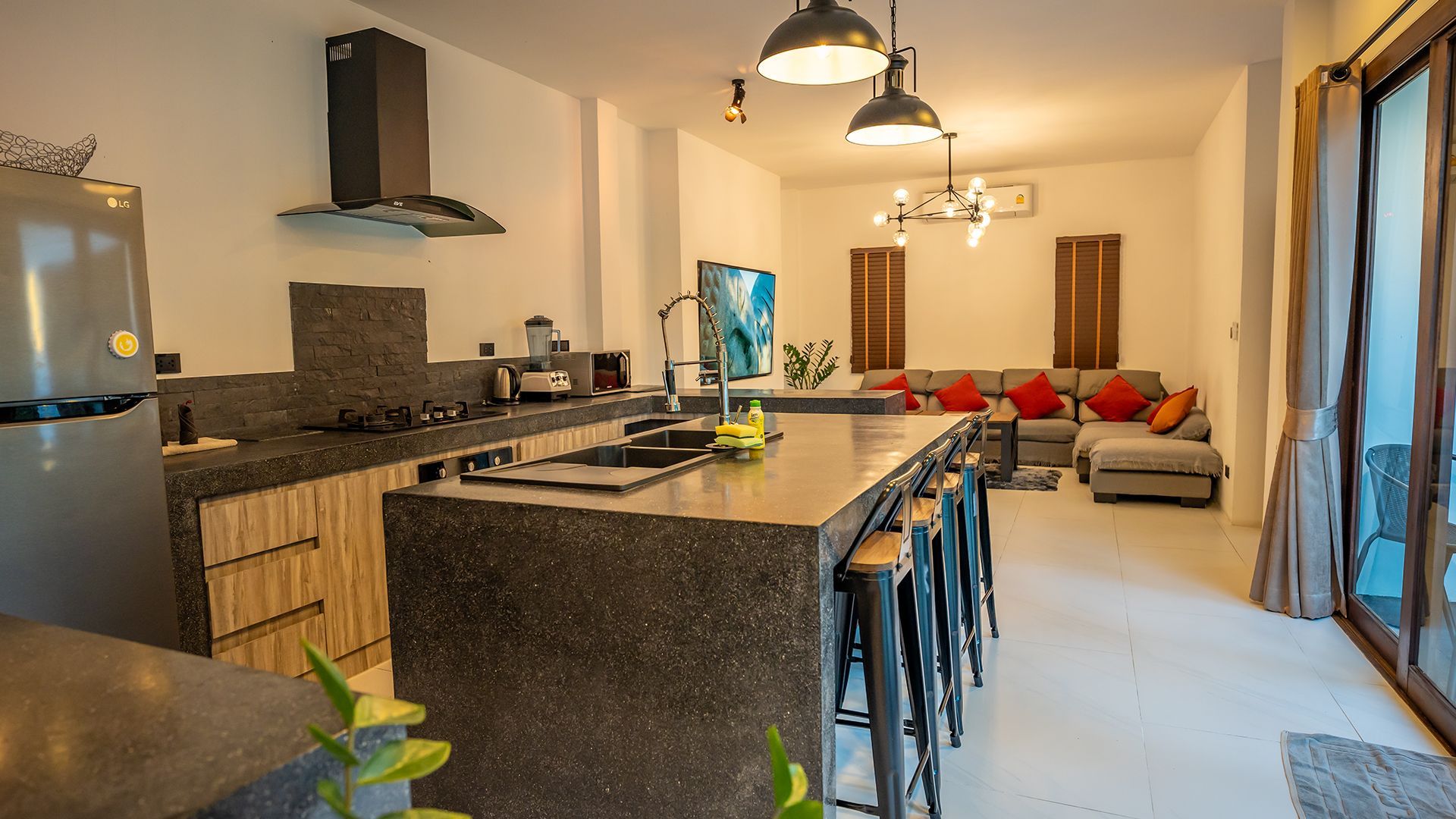 Modern kitchen with granite island and bar stools opening to a cozy living room with sofas and pendant lights