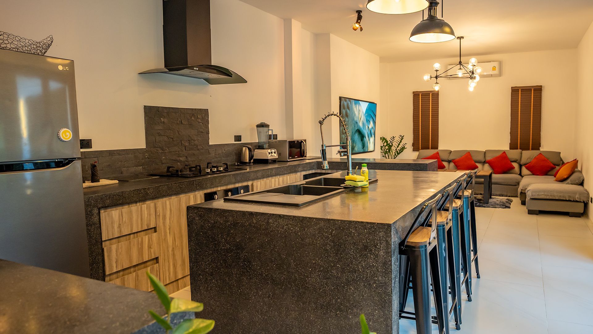Modern kitchen with granite island and stools, opening to a bright living room with red and gray couches.