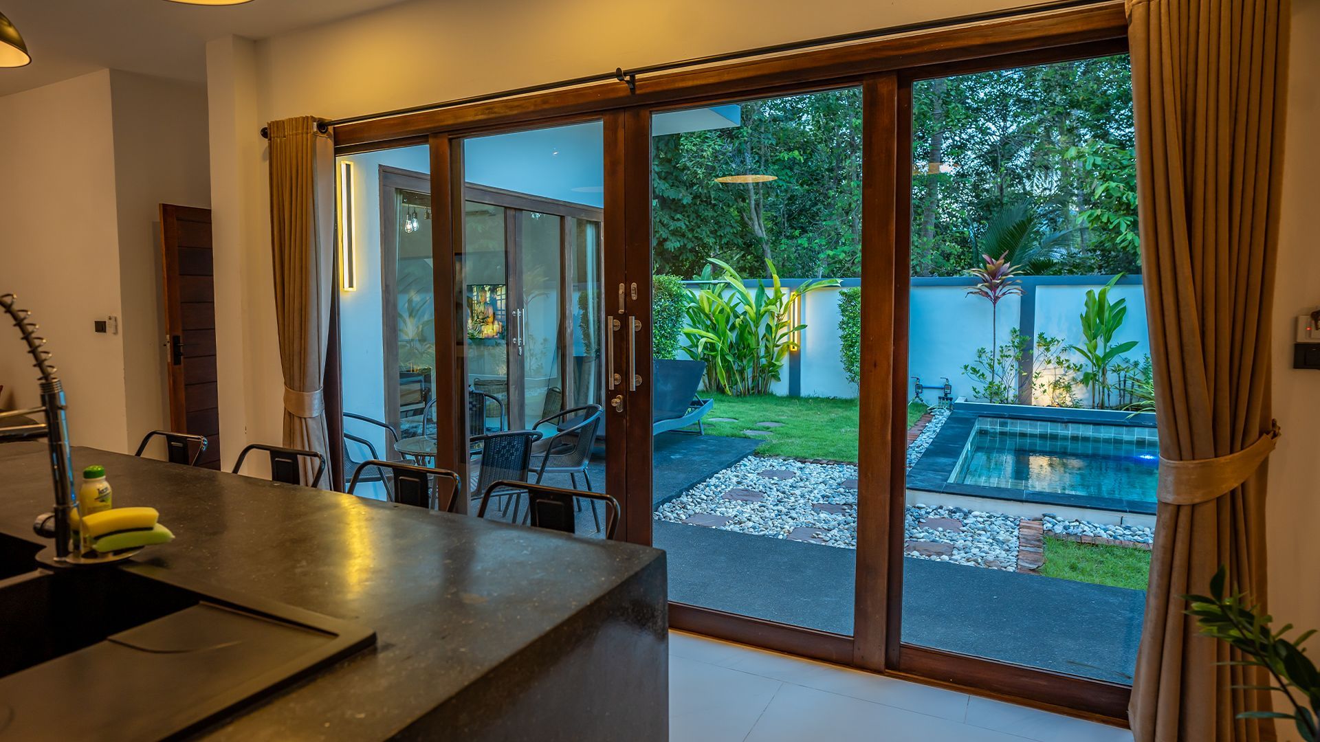 Modern kitchen opening to a patio with sliding glass doors, pool, and lush backyard garden.