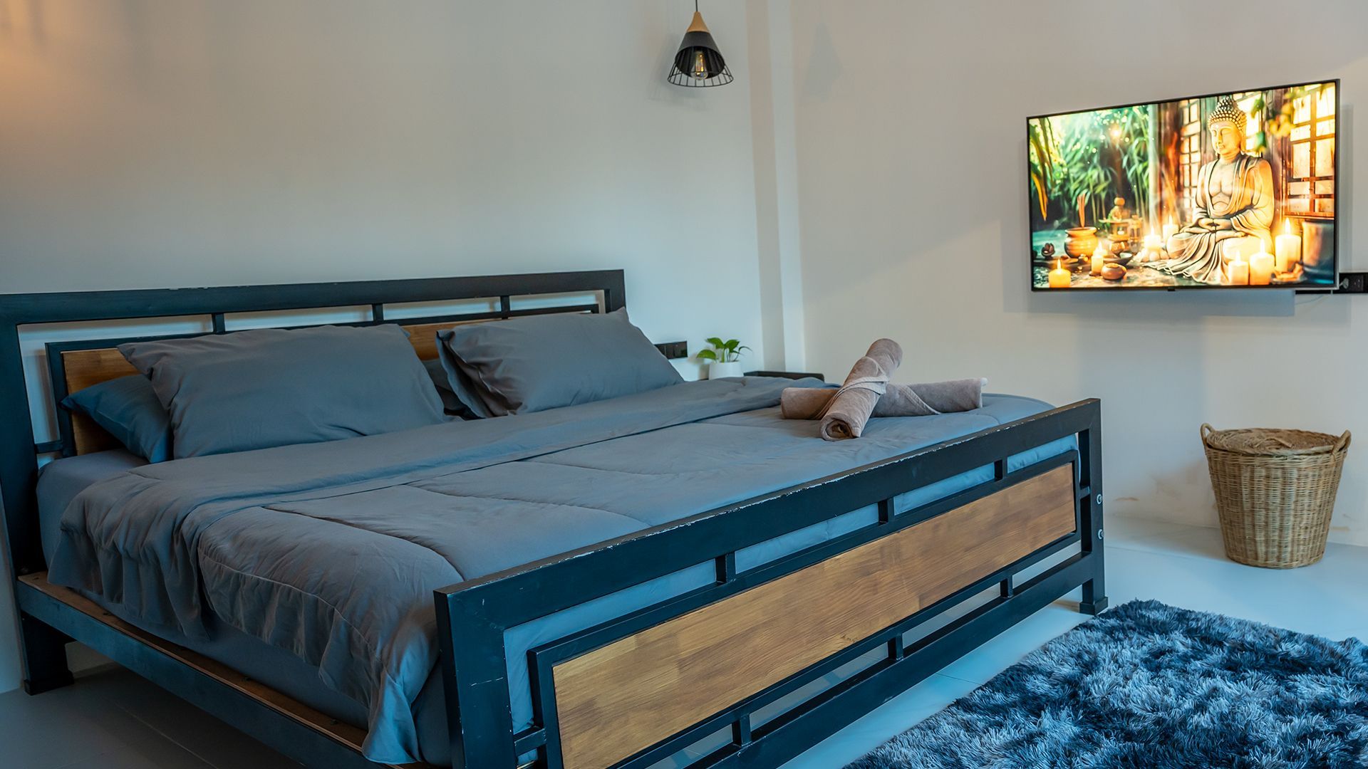 Modern bedroom with a black-framed bed, gray bedding, blue rug, wall-mounted TV, and a wicker basket.