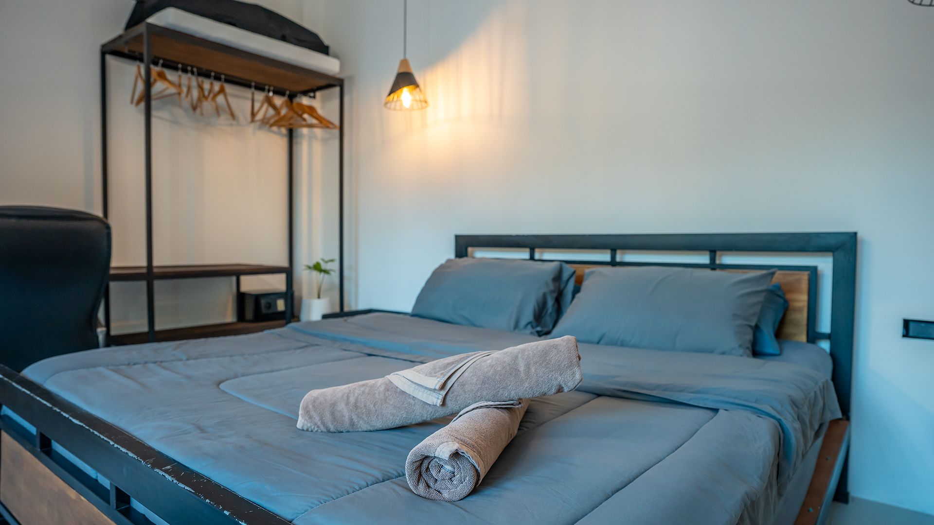Modern bedroom with a gray bed, rolled towels, bedside lamp, and a clothing rack against a white wall