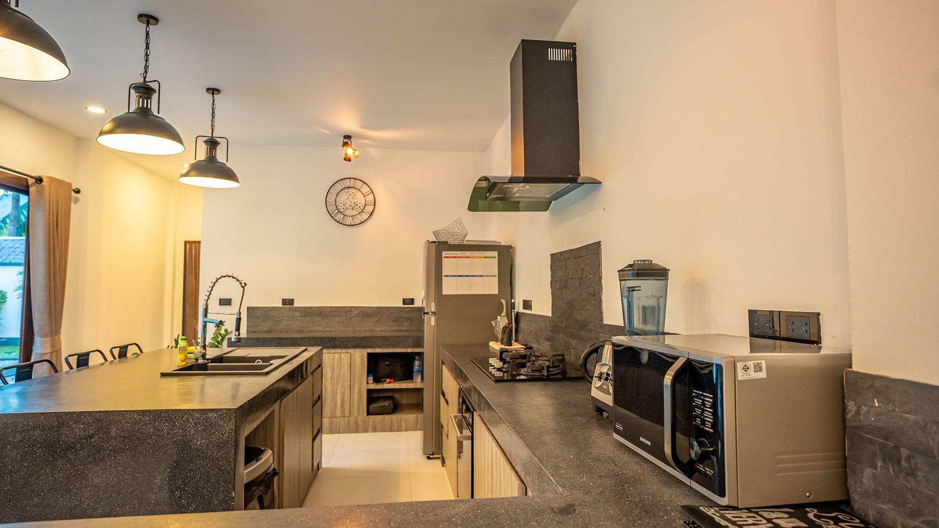Modern kitchen with granite counters, hanging lights, stainless appliances, and a wall clock.