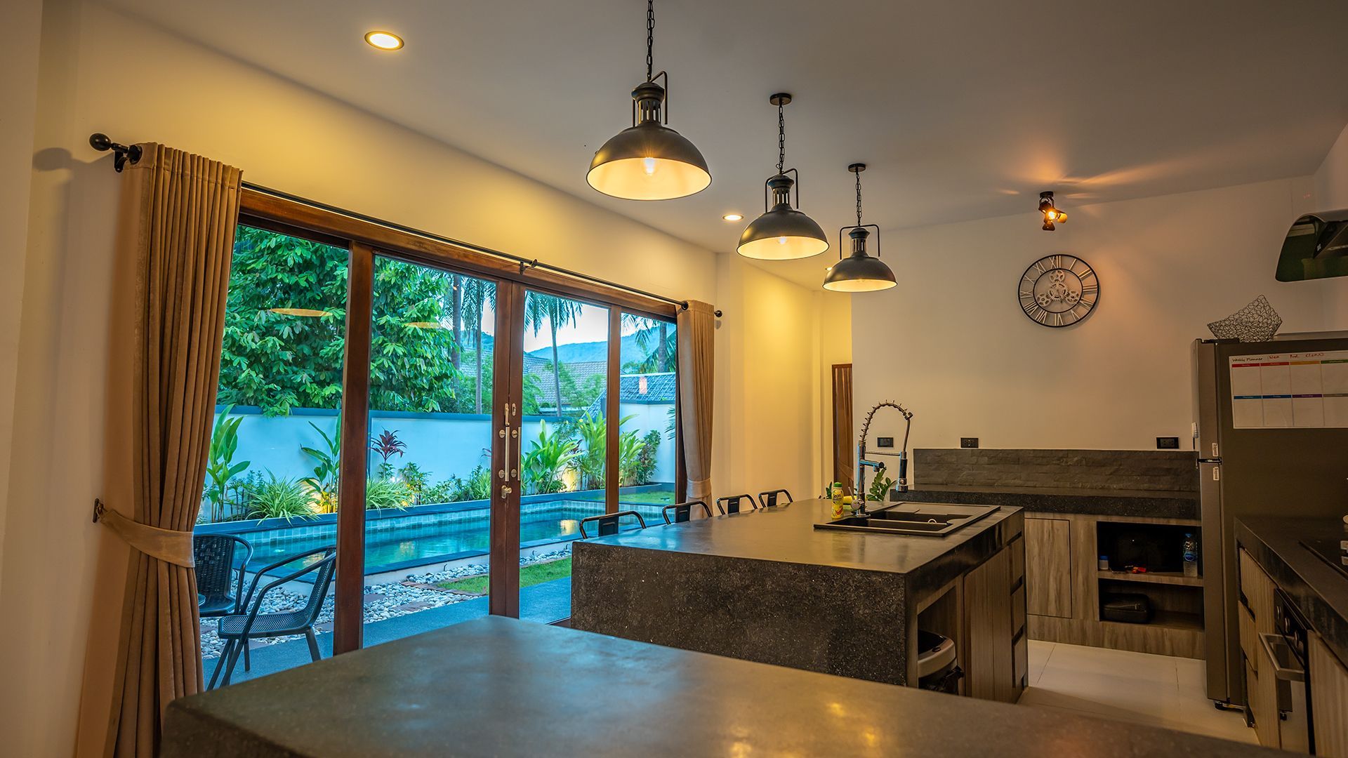 Modern kitchen with island, pendant lights, and large sliding doors opening to a blue-lit patio/garden.