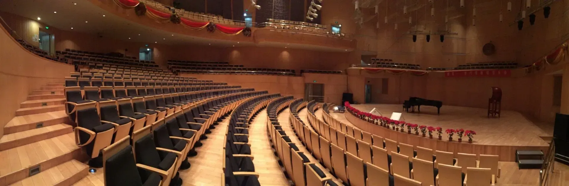 Panoramic view of a modern, curved concert hall with wooden interior, tiered dark seating, and a stage with a piano.