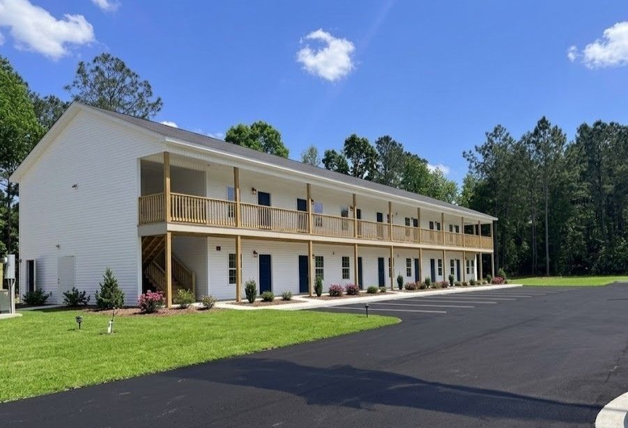 Two-story beige motel with balcony and parking lot under a blue sky, surrounded by trees