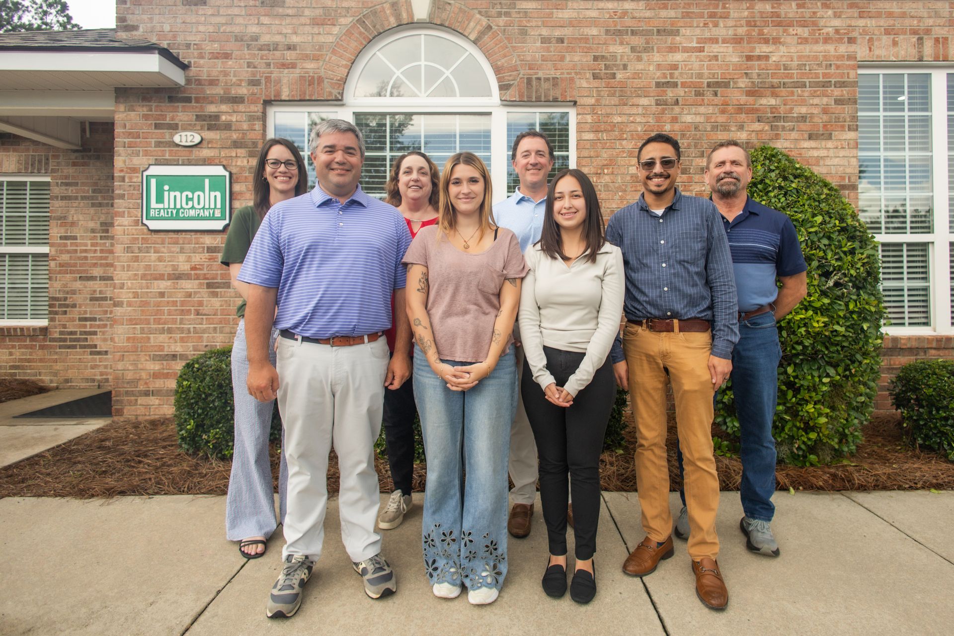 Group of six people standing outside a brick building, smiling in front of the entrance.