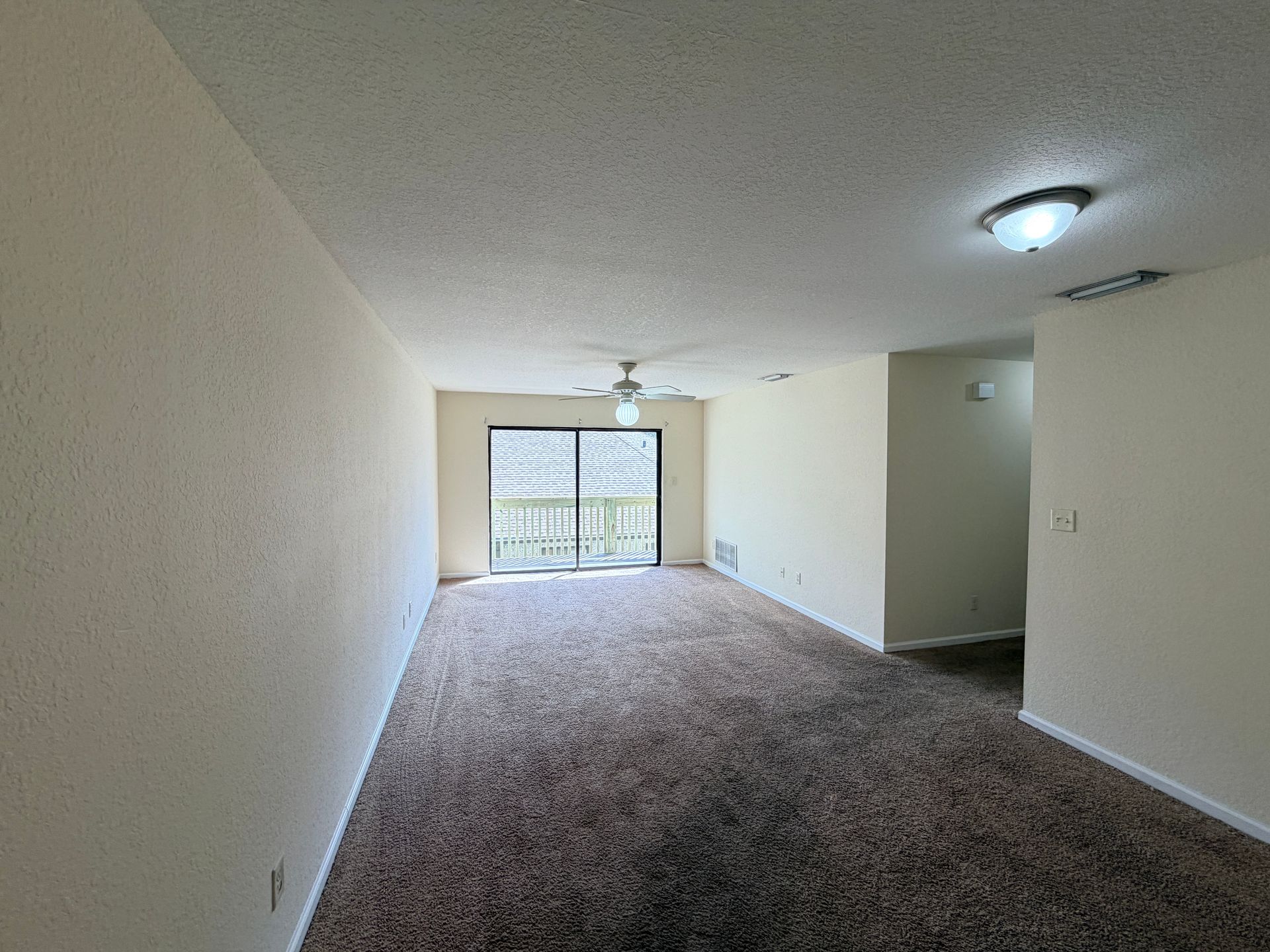 Empty carpeted living room with sliding glass doors and ceiling fan, viewed toward a balcony.