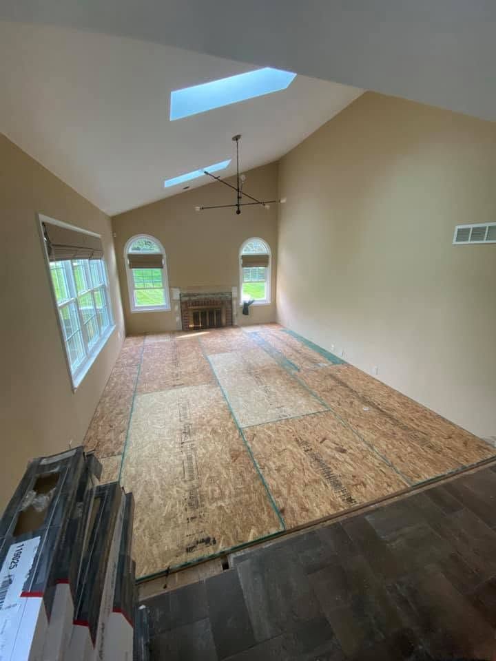 Empty room with vaulted ceiling, skylights, unfinished plywood floor, and windows overlooking greenery.