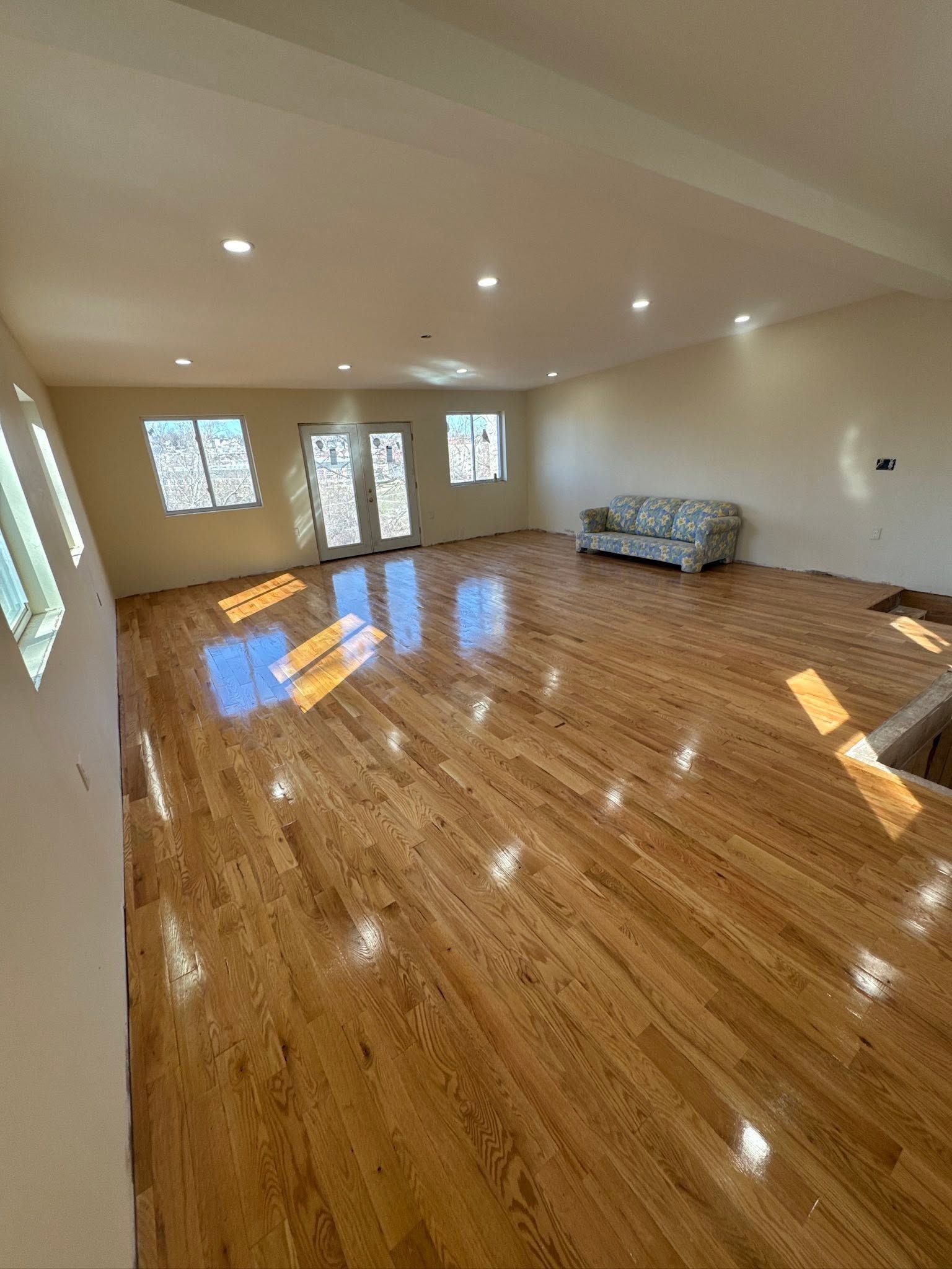 Empty living room with glossy wood floors, recessed lights, windows, and a patterned sofa against the wall.