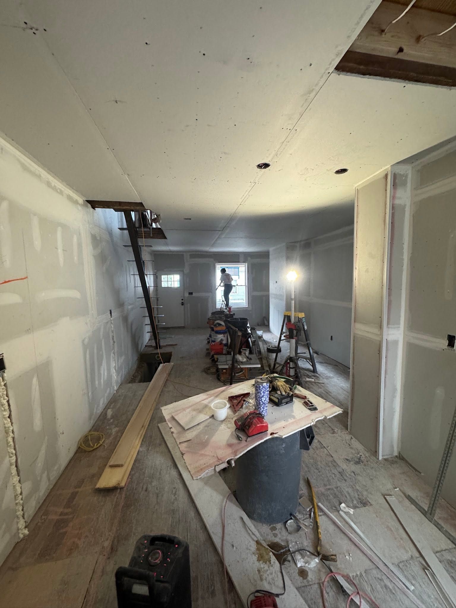 Narrow hallway under renovation with tools, ladders, paint cans, and construction debris on the floor. Narrow hallway under renovation with tools, ladders, paint cans, and construction debris on the floor.