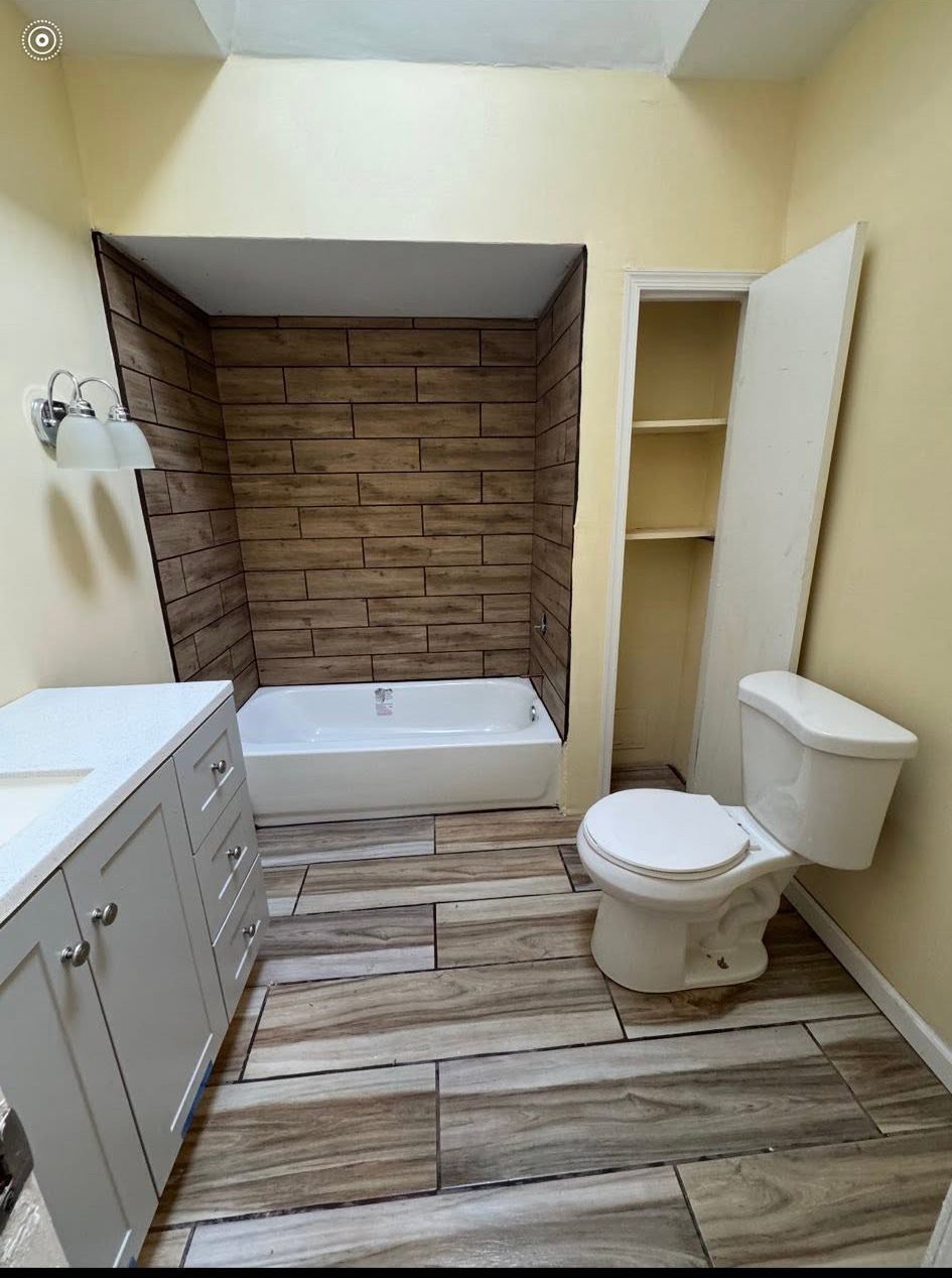 Small bathroom with bathtub, toilet, vanity, wood-look floor, and tiled alcove behind tub. Small bathroom with bathtub, toilet, vanity, wood-look floor, and tiled alcove behind tub.