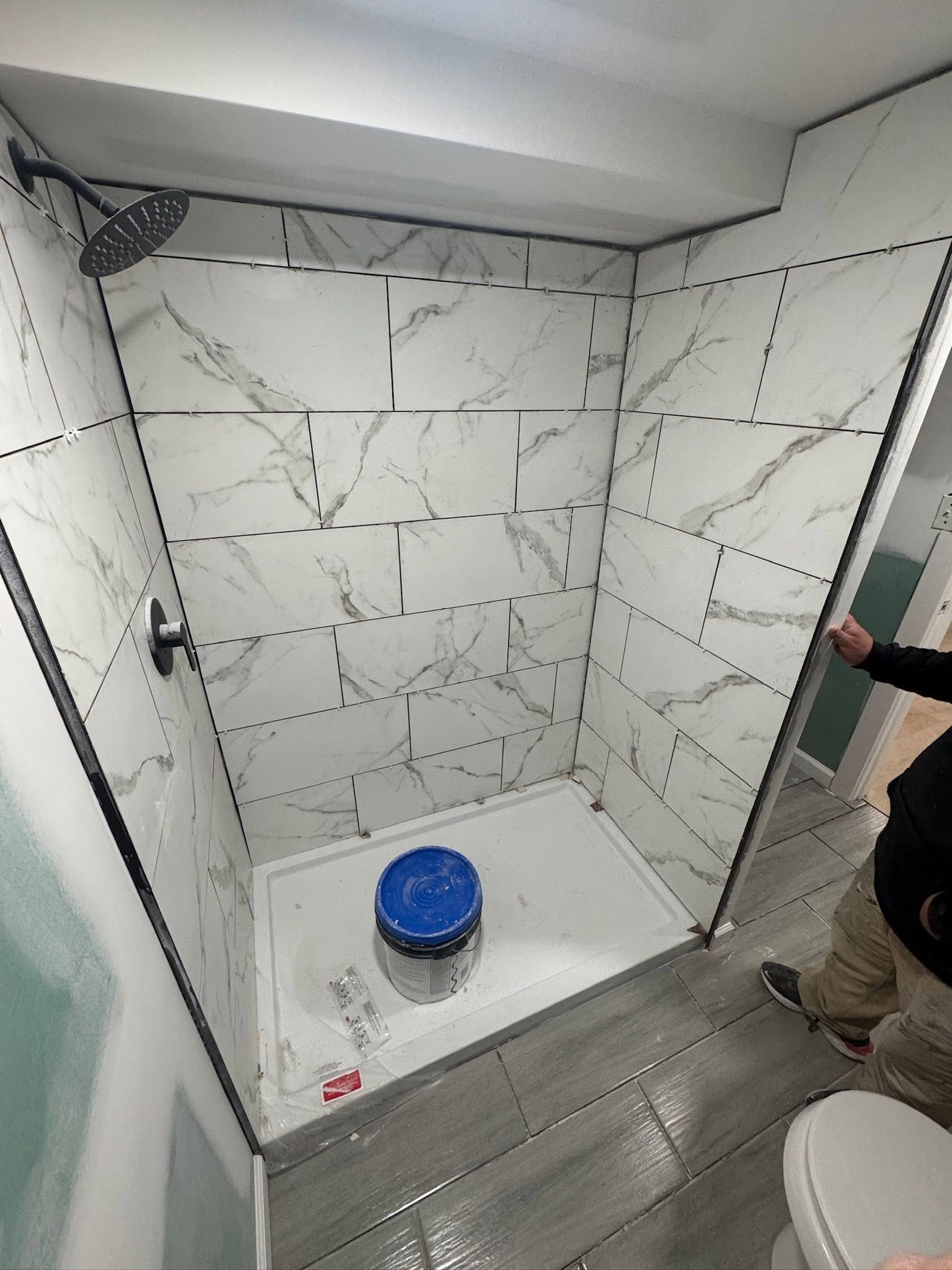 Empty tiled shower stall with a blue bucket on the floor and a partially visible person at the door. Empty tiled shower stall with a blue bucket on the floor and a partially visible person at the door.