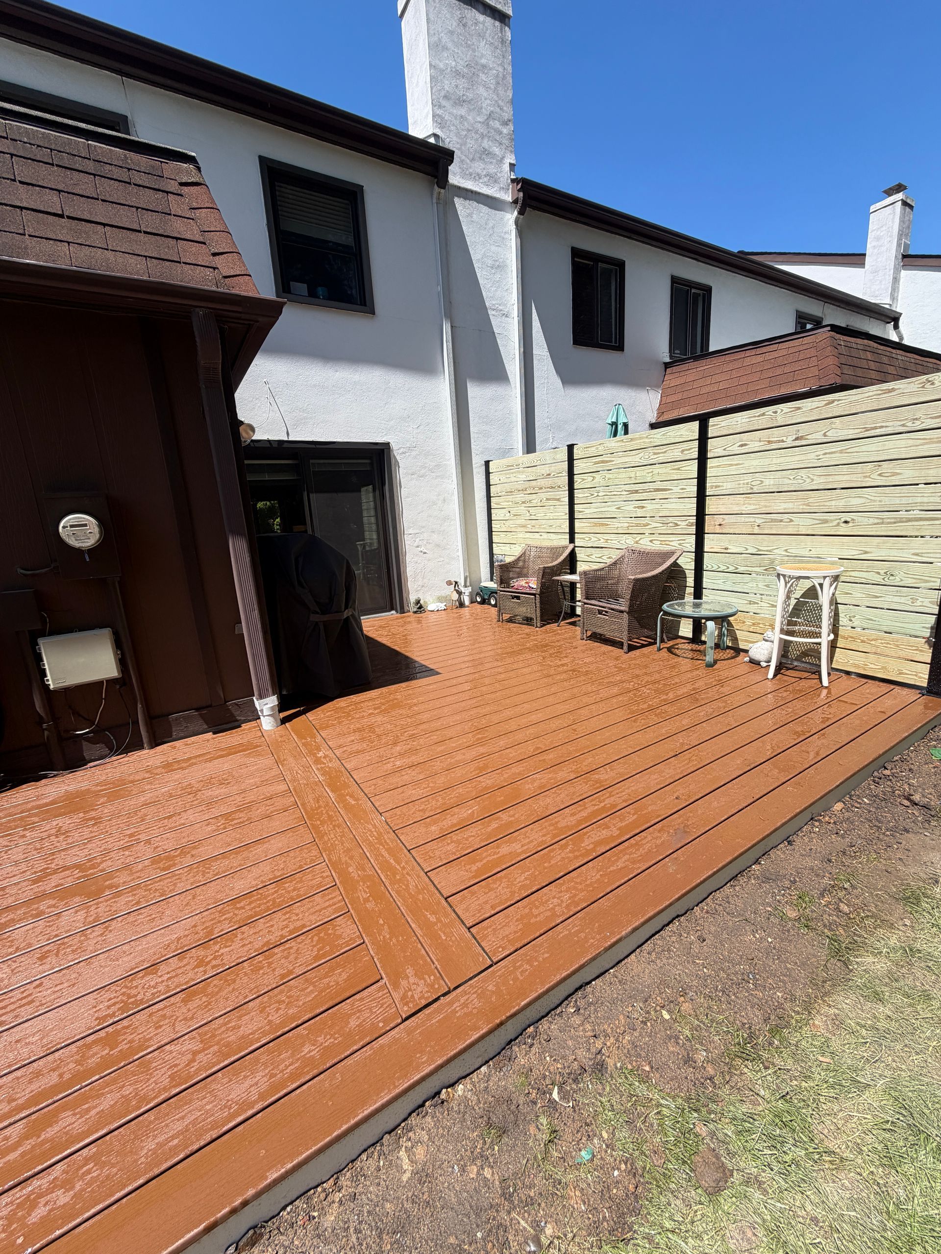 Wooden deck patio beside a white house with stone wall and a paved walkway in sunlight