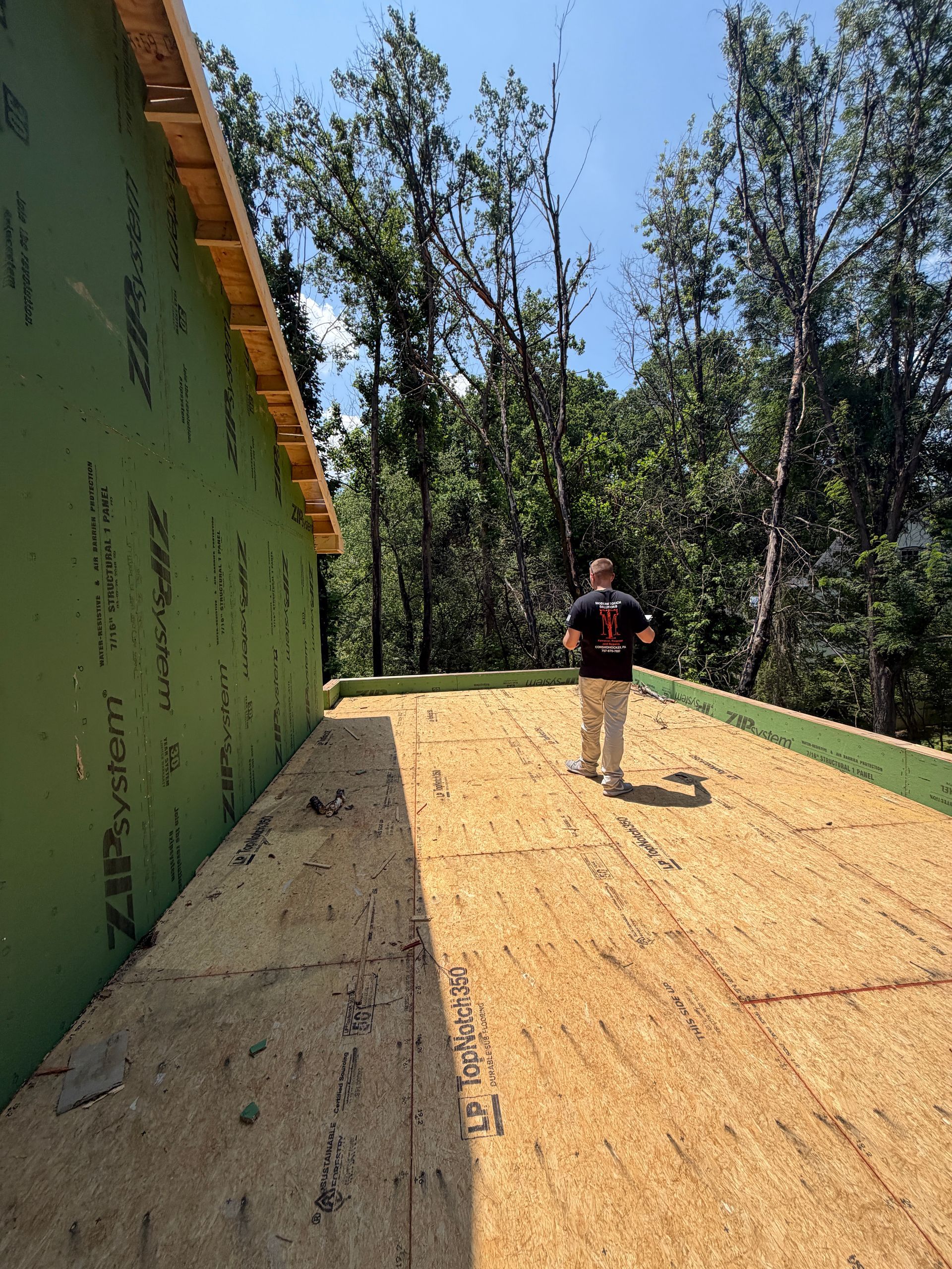 Person standing on a plywood roof next to a green wall, surrounded by trees under a blue sky.