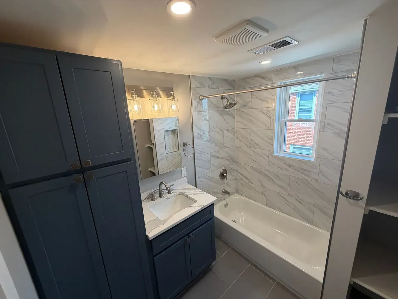 Bright modern bathroom with dark vanity, white tub, marble tile, and a window above the tub.