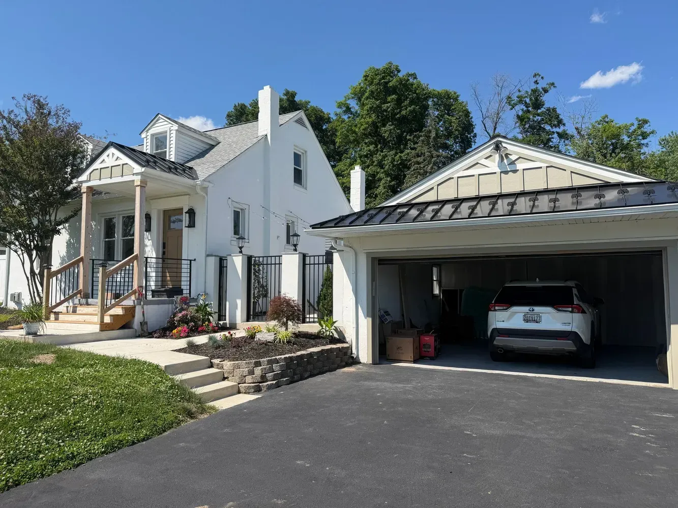 White house with porch and detached garage; car parked in the open garage under a sunny blue sky.