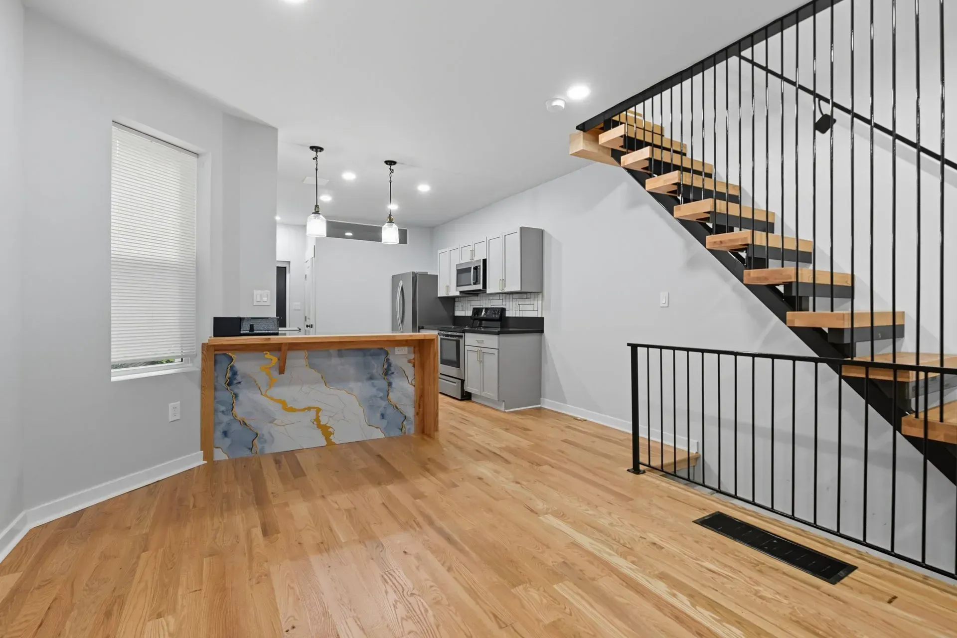 Empty modern living space with hardwood floors, staircase, and compact kitchen with marble island