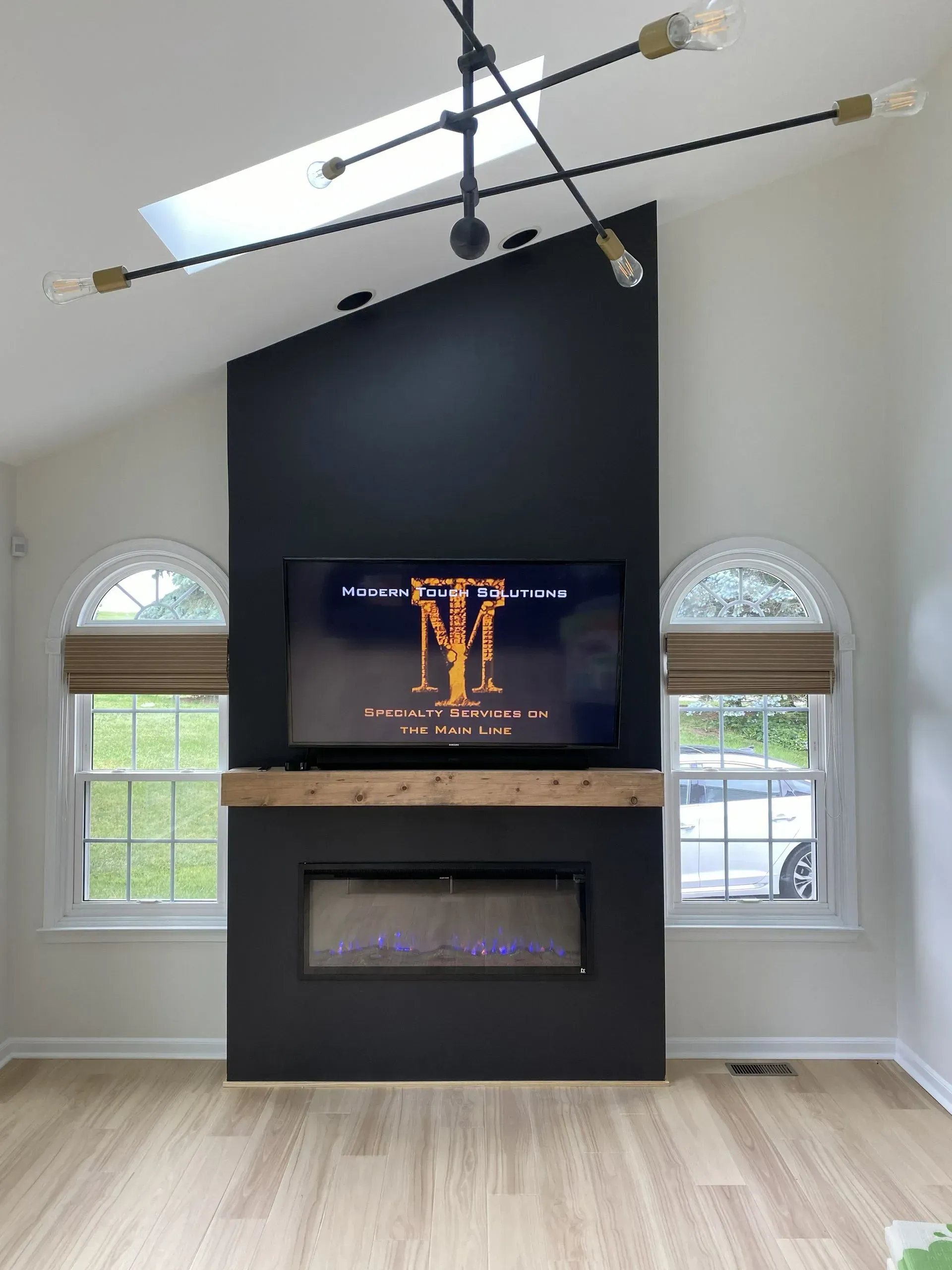 Modern living room with black fireplace wall, TV, arched windows, and ceiling fan above light wood floors