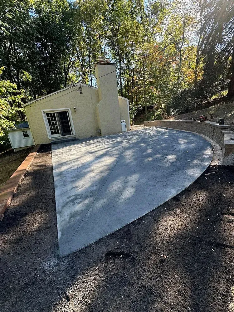 Freshly poured curved concrete patio beside a small beige house surrounded by trees