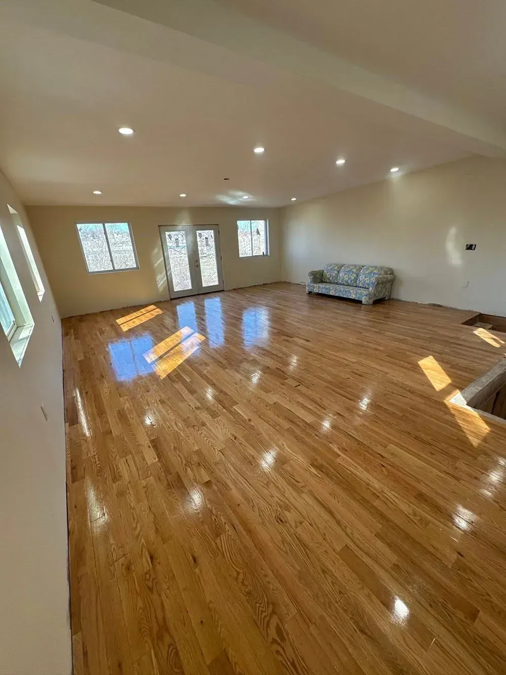 Empty living room with glossy hardwood floors, white walls, recessed lights, and a gray sofa against the far wall