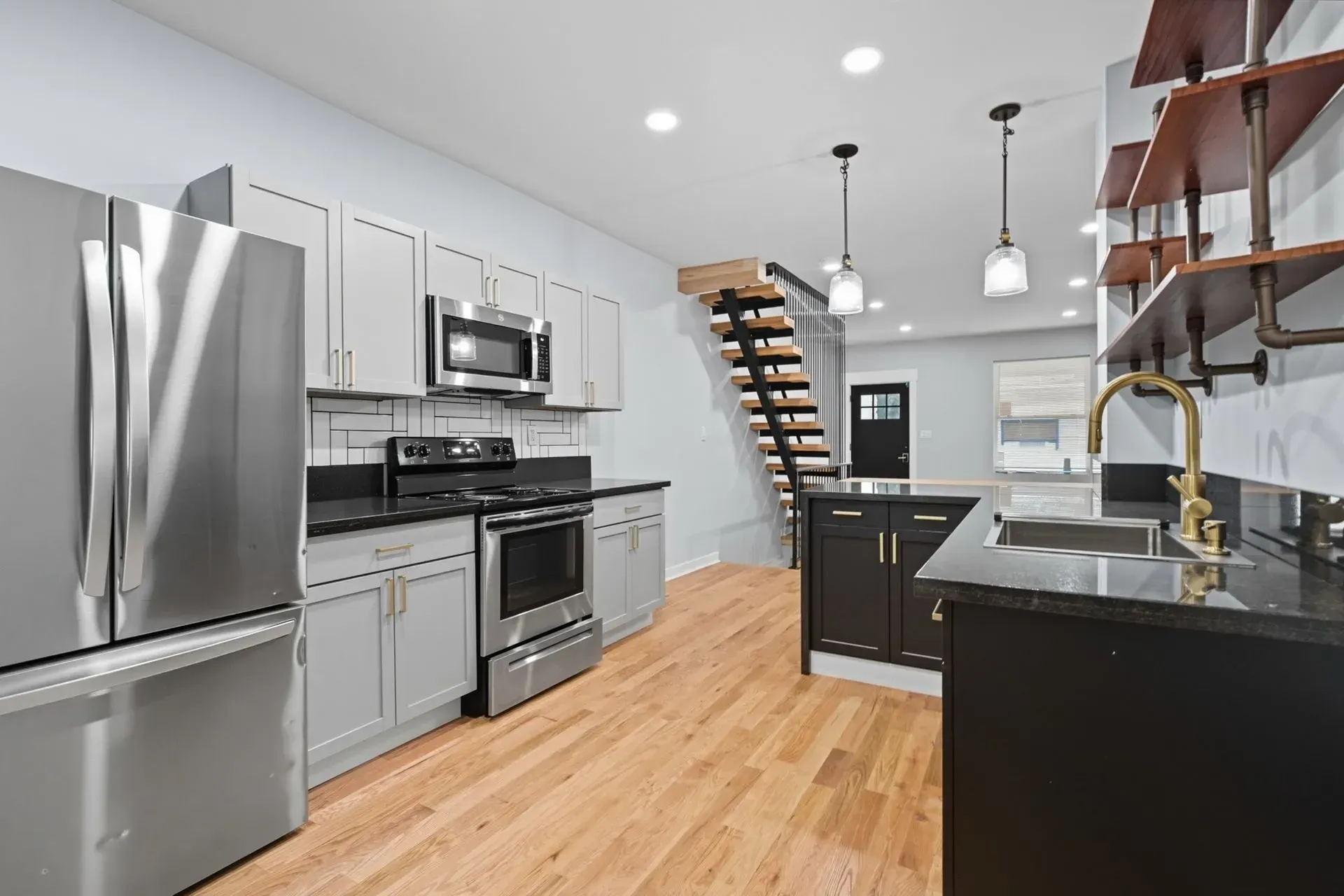 Modern kitchen with stainless steel appliances, dark cabinets, wood floors, and a staircase to an upper level