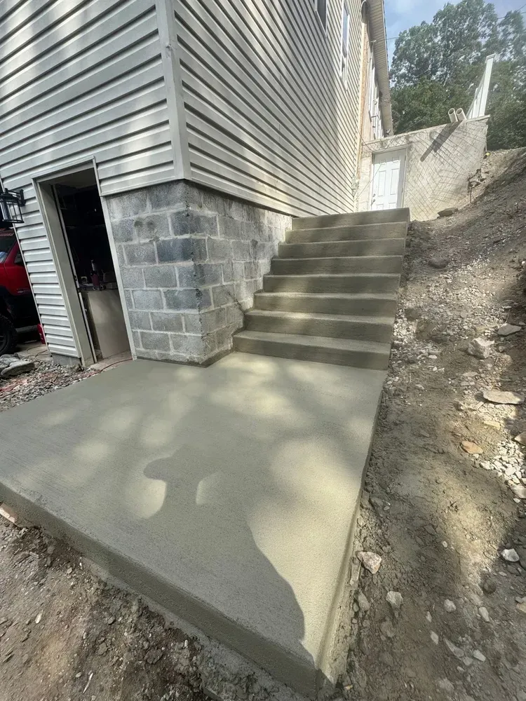 Concrete stairs beside a house, leading up a dirt slope, with a freshly poured patio below