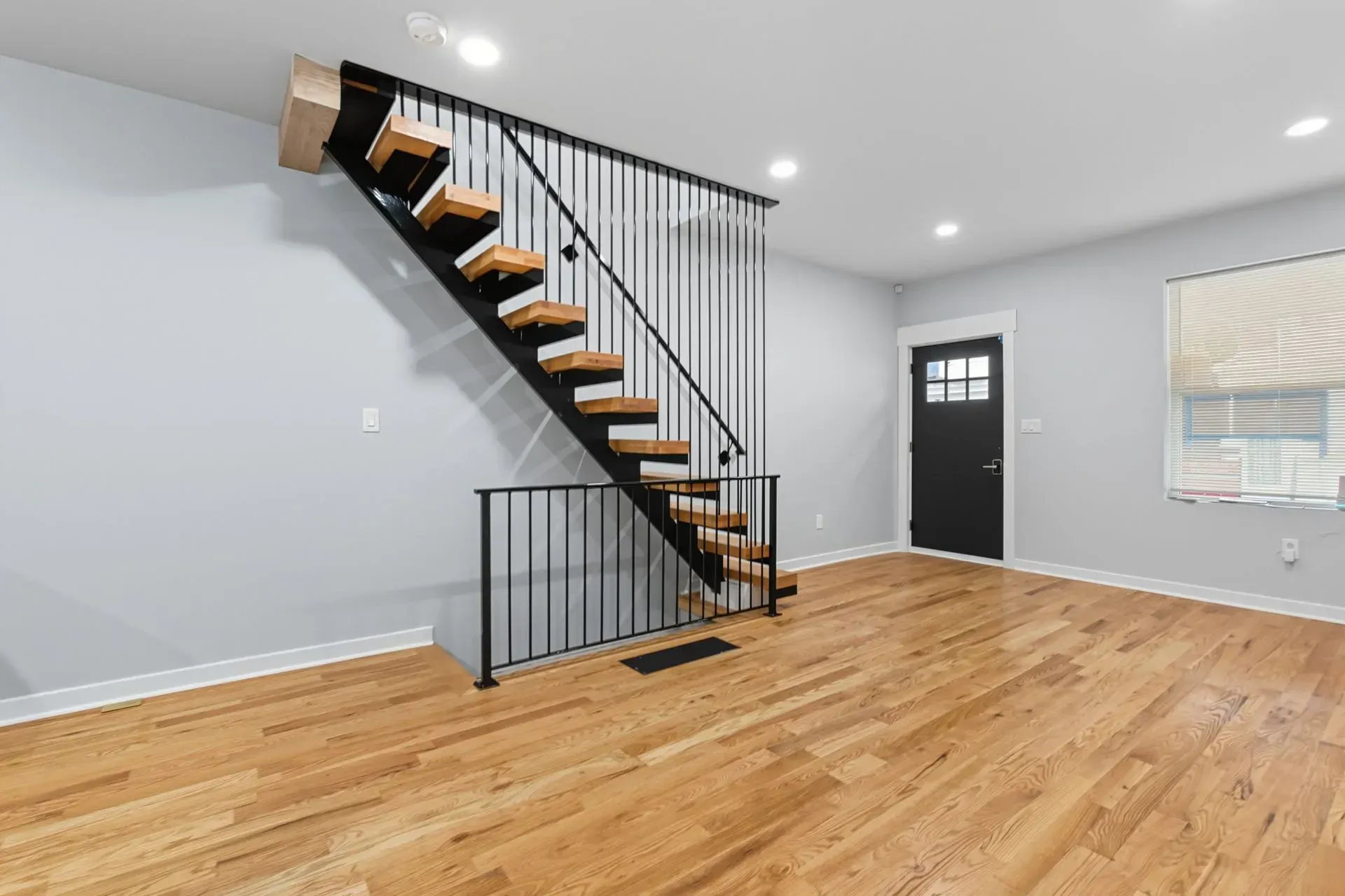 Empty room with hardwood floors and black metal staircase, gray walls, and a window with blinds.