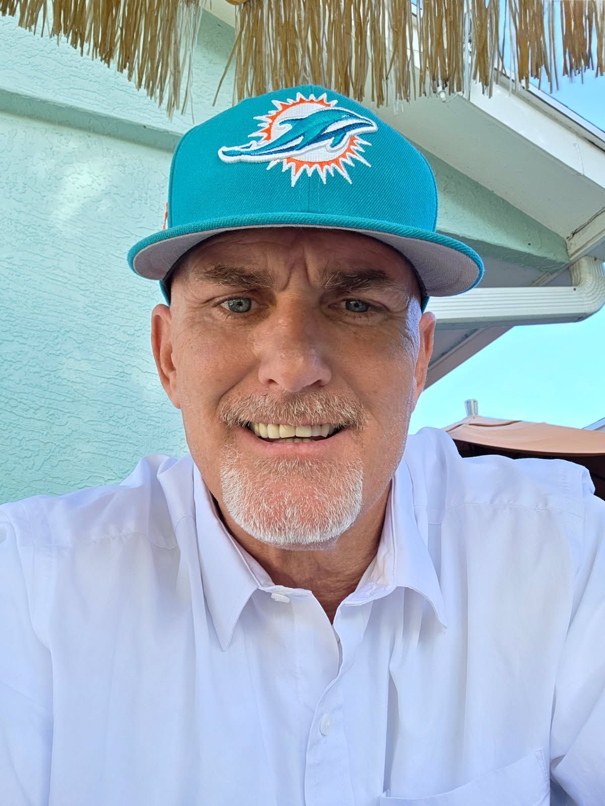 Man wearing a Miami Dolphins hat, smiling, white shirt, outdoors.