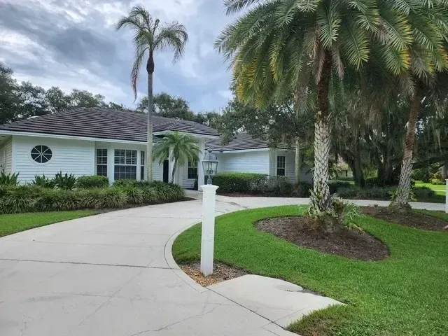 White house with curved driveway, palm trees, and green grass on a cloudy day.
