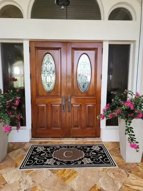 Double wooden front doors with oval glass panes, flanked by windows and flower pots, over a welcome mat.
