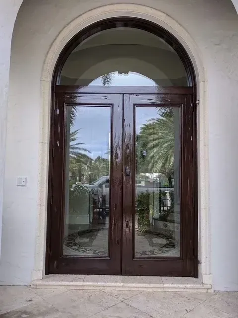 Dark wood double doors with arched glass window, framed in white, reflecting palm trees.
