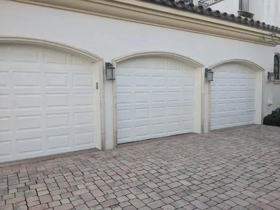 Three white garage doors with arched tops, flanked by lights, set on a brick driveway.