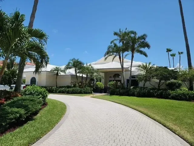 Paved driveway leads to a white building with tropical landscaping and palm trees under a blue sky.