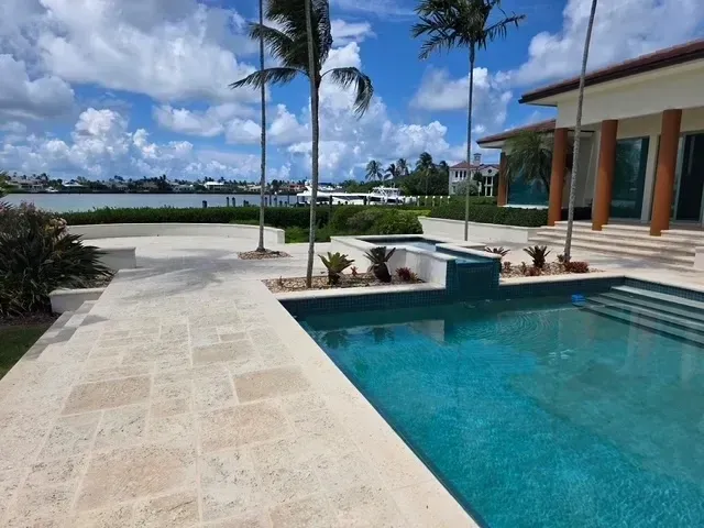 Luxury home with pool, patio, palm trees, and waterfront view under a blue, cloudy sky.
