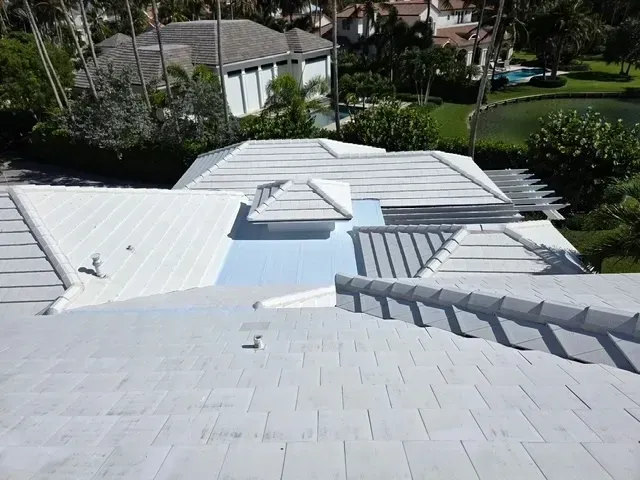 White roof of a home with multiple sections, trees, and a pond visible in the background.