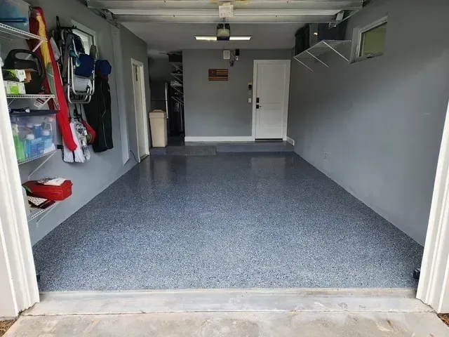 A garage interior with a gray epoxy floor. Walls are painted gray. Shelves with items are on the left. The garage door is open.