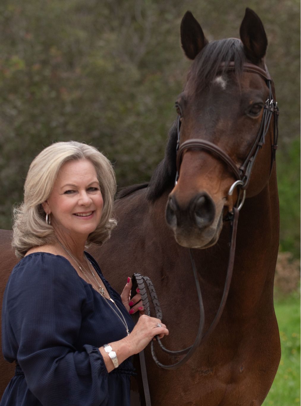 Woman with blonde hair smiles next to a brown horse in an outdoor setting.