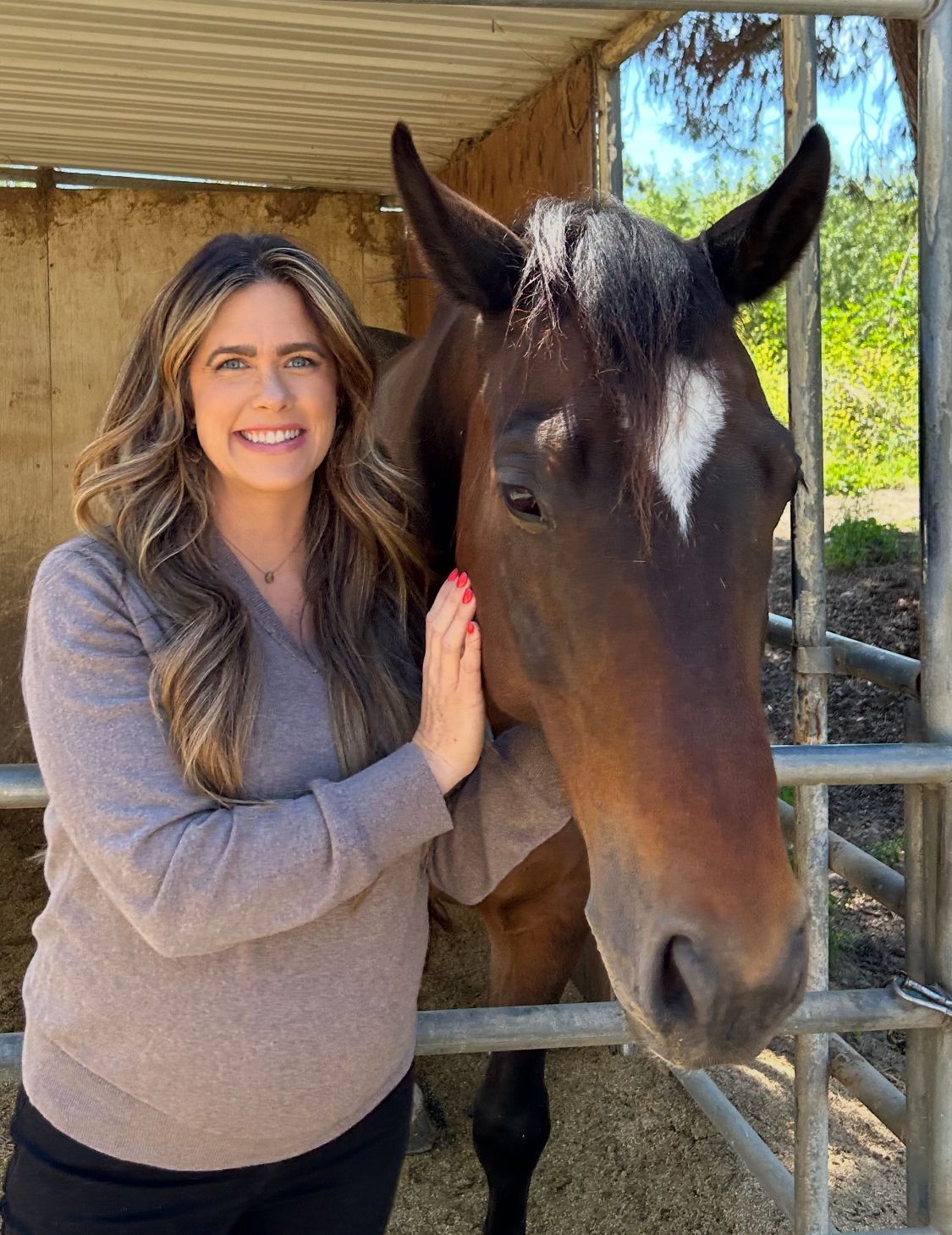 Woman in gray sweater petting a brown horse in a stable; they are smiling.