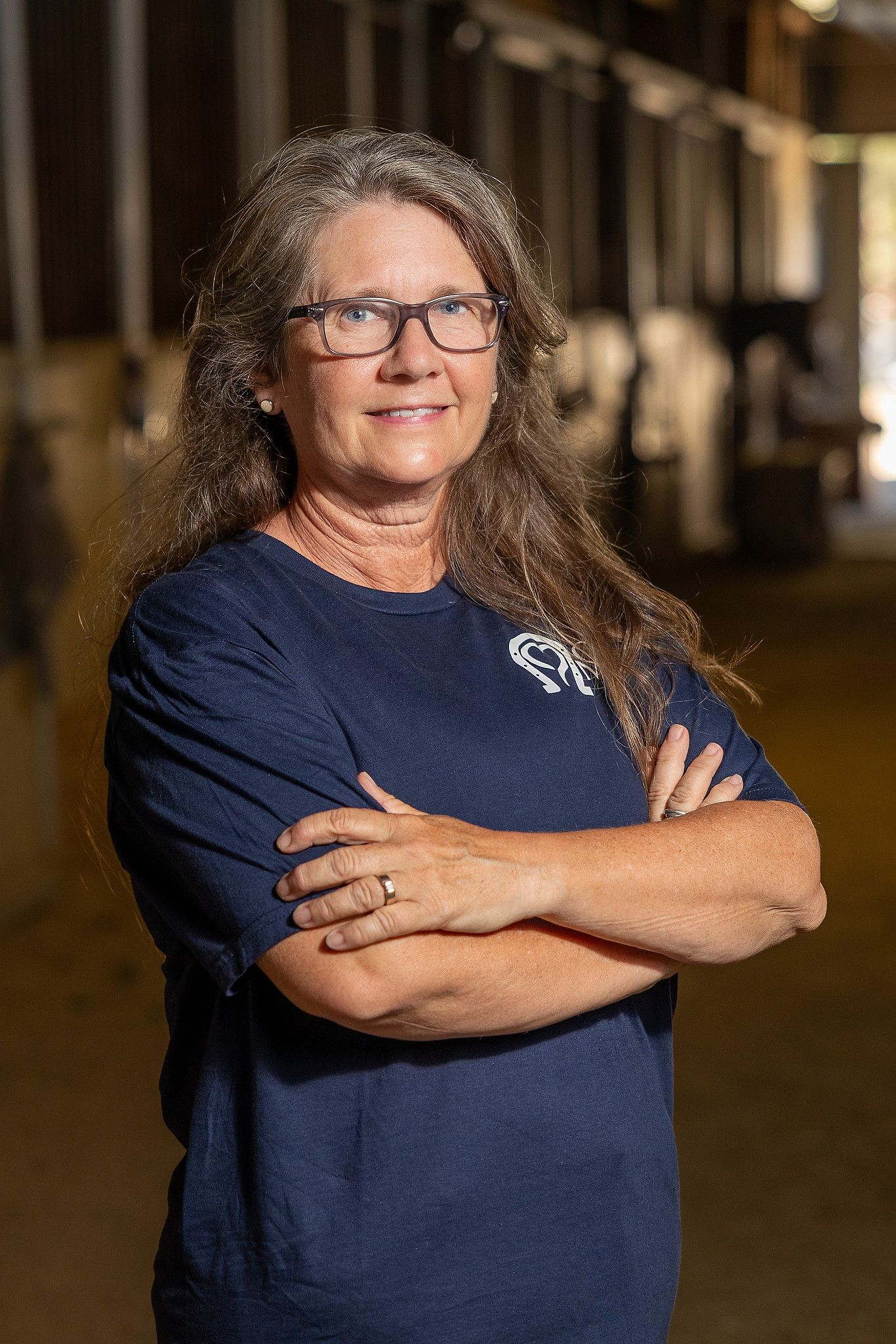 Woman in blue shirt stands beside a chestnut horse, under a tree.