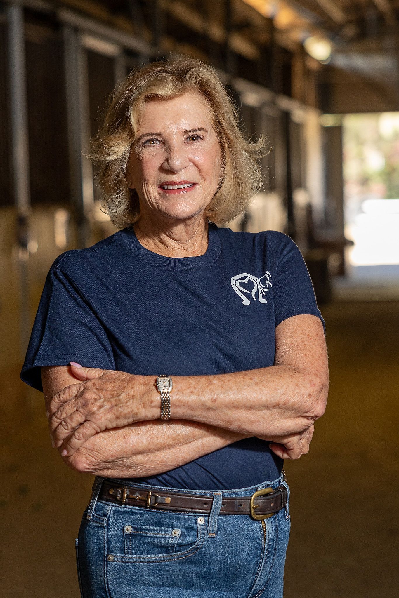 Woman standing with arms crossed in a blue T-shirt and jeans in a dim indoor hallway.