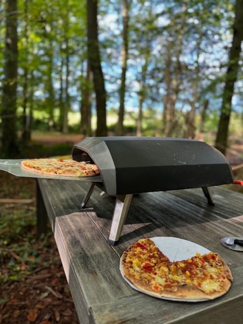 Pizza oven cooking pizza on a wooden table in a forest. One pizza is in the oven, another is sliced nearby.